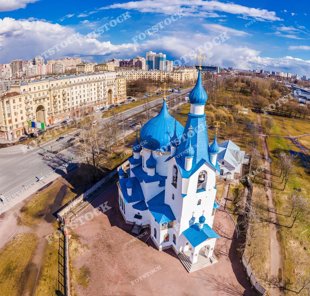 На переднем плане изображен православный храм с характерными голубыми куполами, символизирующими чистоту и небесную благодать. Здание храма гармонично сочетается с окружающими многоэтажными жилыми домами, создавая контраст между древними традициями и современным Петербургом.